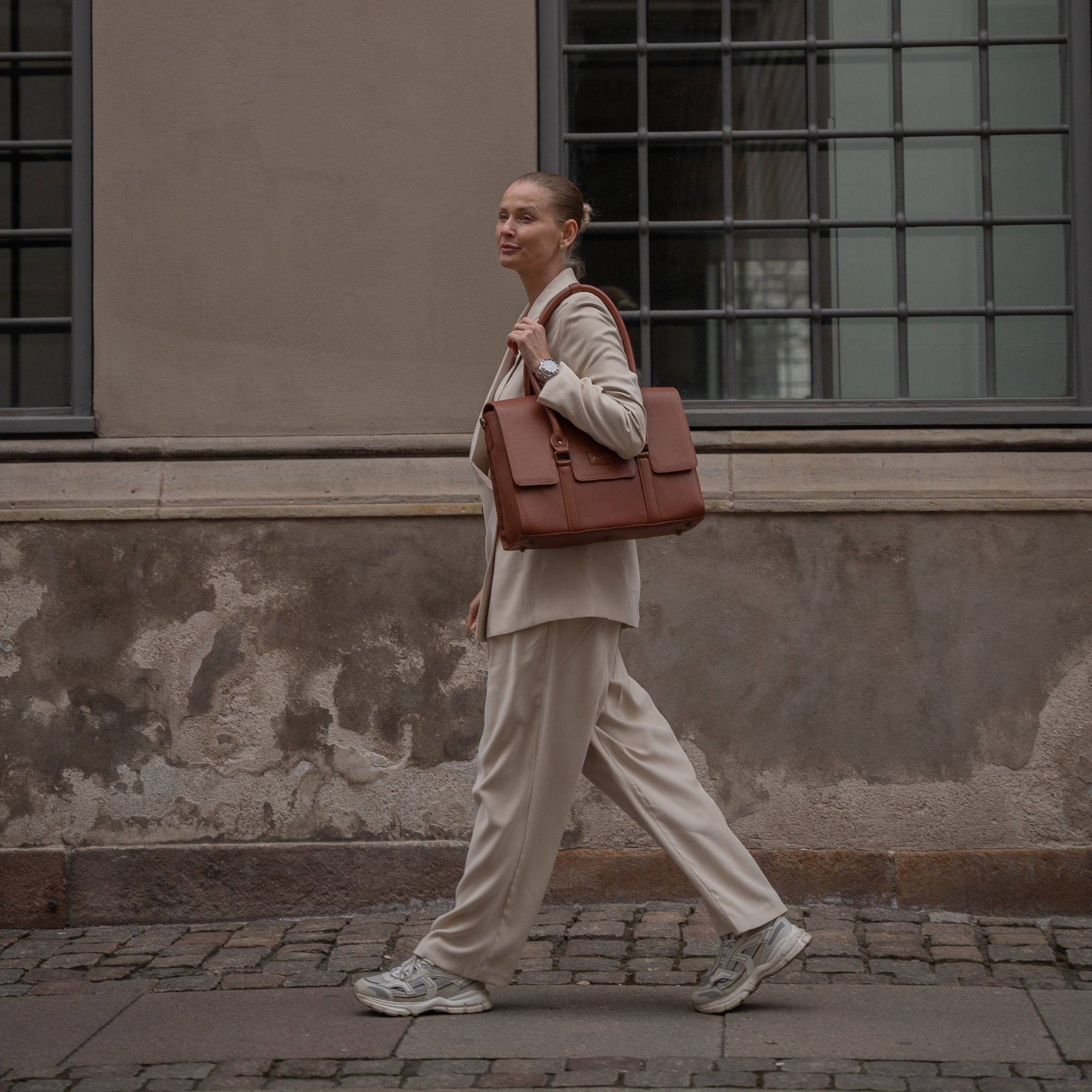 CC Arbejdstaske i Cognac læder over skulderen på gående kvinde i et lyst oversized jakkesæt