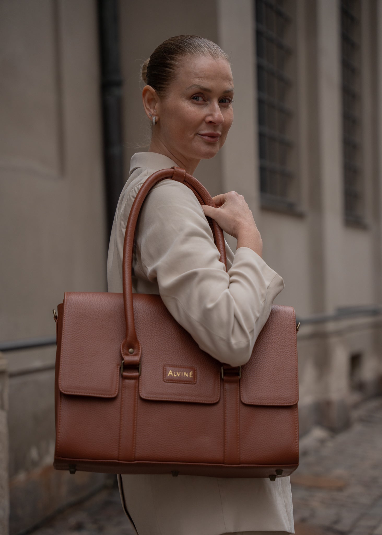 CC Arbejdstaske i Cognac læder over skulderen hos kvinde i et lyst oversized jakkesæt der kigger smilende ind i kameraet