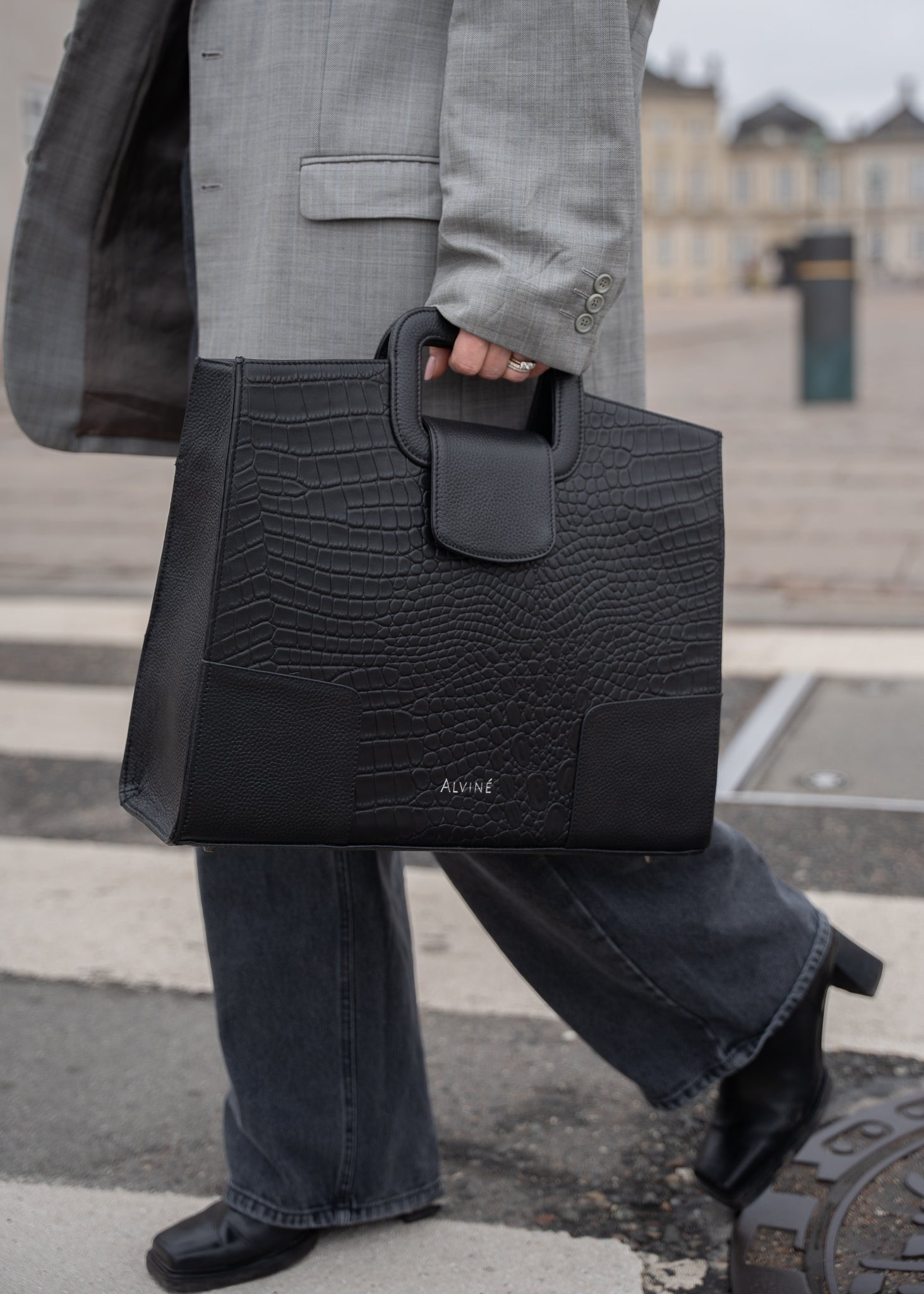Close up af VGC Arbejdstaske i sort læder med krokodillestruktur i hånden på gående kvinde i en oversize blazer.