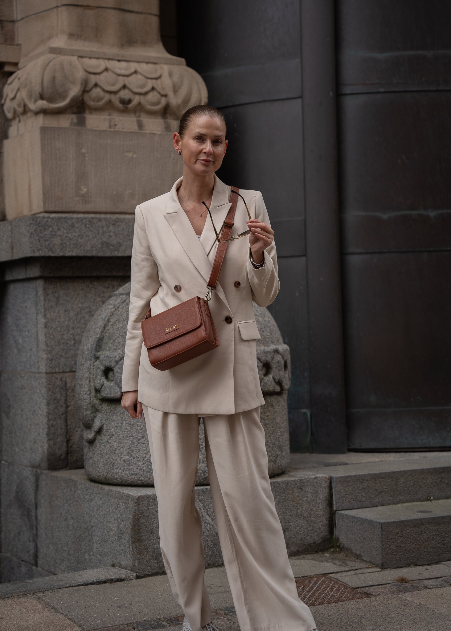 CC Clutch i Cognac læder som crossbody på kvinde i lyst jakkesæt der står med solbriller i hånden og kigger ind i kameraet