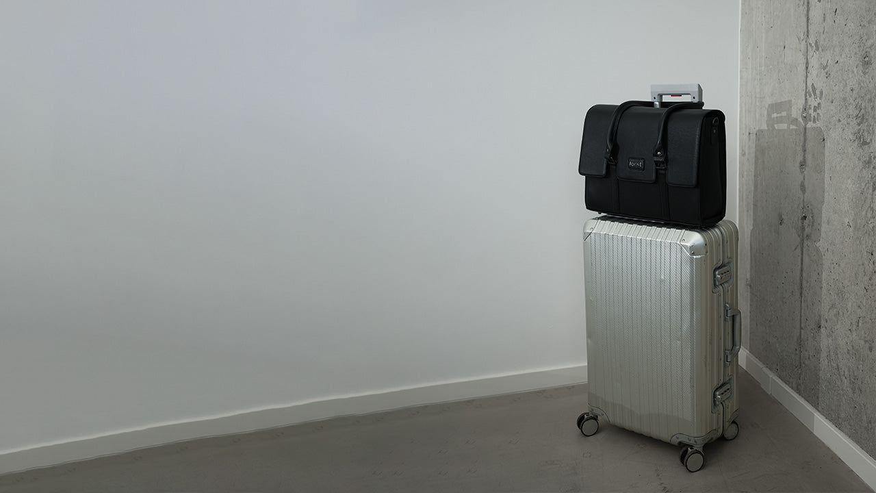 CC Arbejdstaske i sort læder der står på en aluminiums kuffert med håndtagene over trolley håndtaget